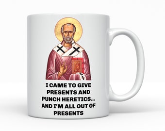 May include: White ceramic mug featuring an image of a religious figure with a gold halo and the text "I CAME TO GIVE PRESENTS AND PUNCH HERETICS... AND I'M ALL OUT OF PRESENTS". The mug has a white handle.