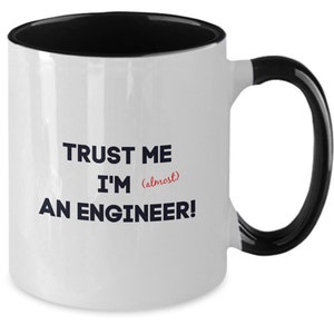 May include: White ceramic mug with a black rim and handle. The mug has the text "TRUST ME I'M (almost) AN ENGINEER!" printed on it in black and red.