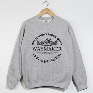 May include: Gray sweatshirt with a black graphic design featuring a mountain range, a sun, and trees. The text reads "Miracle Worker Promise Keeper Waymaker - Isaiah 42:16 - Light in the Darkness".