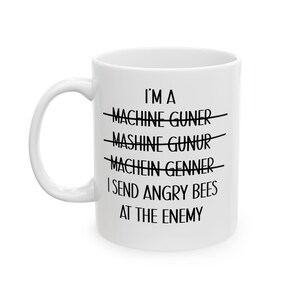 May include: White ceramic mug with the text "I'M A MACHINE GUNER, MASHINE GUNUR, MACHEIN GENNER, I SEND ANGRY BEES AT THE ENEMY" in black. The text is crossed out multiple times. The mug has a white handle.