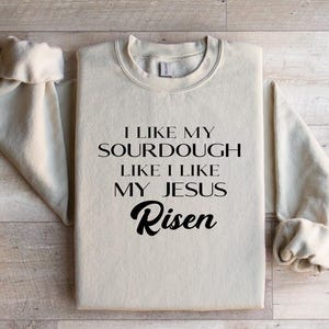 Peut inclure: Un sweat-shirt beige avec le texte "J'aime mon levain comme j'aime mon Jésus Ressuscité" imprimé en noir.