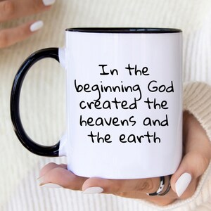 May include: White ceramic mug with a black handle and rim. The mug features the text "In the beginning God created the heavens and the earth" in black script. The mug is held by a person.
