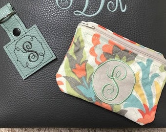 May include: A small, floral zippered pouch with a circle monogram of the letter "S" in green thread. The pouch is on a black textured surface. There is also a small, square, teal leather keychain with a circle monogram of the letter "S" in black thread.