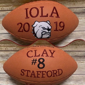 Op de afbeelding: Twee bruine leren voetballen. De bovenste voetbal heeft de tekst "IOLA 20 19" en een wit bulldog-mascotte. De onderste voetbal heeft de tekst "CLAY #8 STAFFORD".