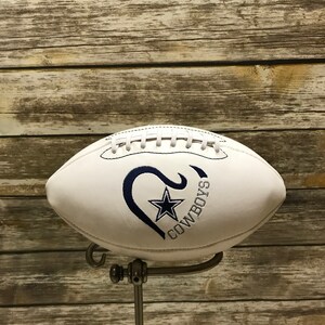 Op de afbeelding: Een witte voetbal met een blauw geborduurd hart en het woord "COWBOYS" erop. De voetbal staat op een zilveren standaard.