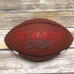 Op de afbeelding: Een bruine leren voetbal met witte strepen en de tekst "RICHARDS 2018" geborduurd in rood en grijs.