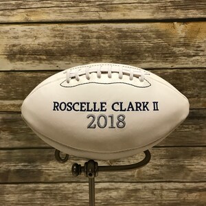 Op de afbeelding: Een witte leren voetbal met blauwe borduurwerk met de tekst "Roscelle Clark II 2018" op een metalen standaard.