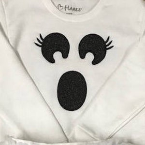 Pode incluir: Camiseta branca de manga comprida com um design de rosto de fantasma preto com glitter. O fantasma tem dois olhos com cílios e uma boca grande.
