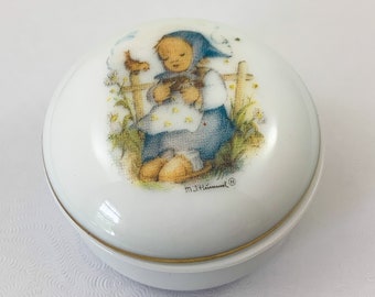 May include: White porcelain trinket box with a gold trim. The lid features a colorful illustration of a young girl holding a small animal. The illustration is signed "M.J.Hummel" with a registered trademark symbol.