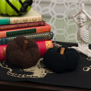 May include: Two knitted pumpkins, one brown and one black, with cinnamon sticks as stems, are displayed on a black fabric with a skull design. A stack of books with titles like "Midsummer Night's Dream" and "Brave New World" are in the background, along with a white skeleton figurine in a yoga pose.