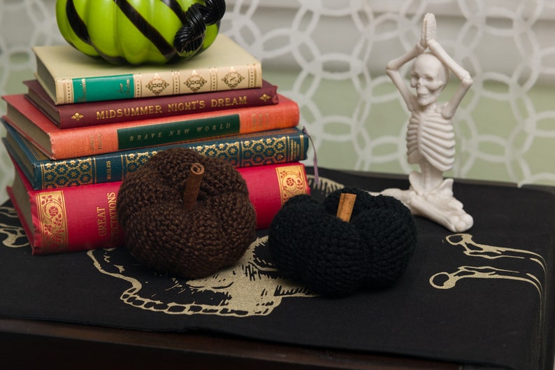 May include: Two knitted pumpkins, one brown and one black, with cinnamon sticks as stems, are displayed on a black fabric with a skull design. A stack of books with titles like "Midsummer Night's Dream" and "Brave New World" are in the background, along with a white skeleton figurine in a yoga pose.