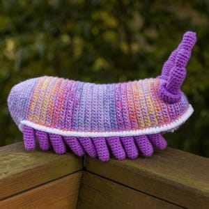 May include: A crocheted purple and pink plush toy shaped like a segmented worm with a white crocheted border and a long, pointed tail.