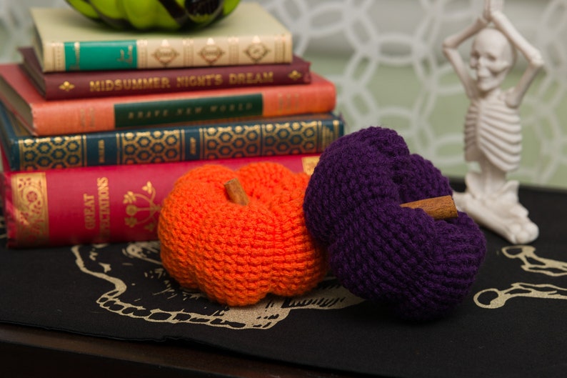 May include: Two crocheted pumpkins, one orange and one purple, with cinnamon sticks for stems. The pumpkins are sitting on a black surface with a skull and crossbones design.