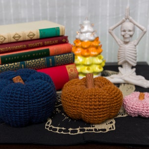May include: Three crocheted pumpkins in different colors: navy blue, brown, and pink. The pumpkins are sitting on a black surface with a white skull design.