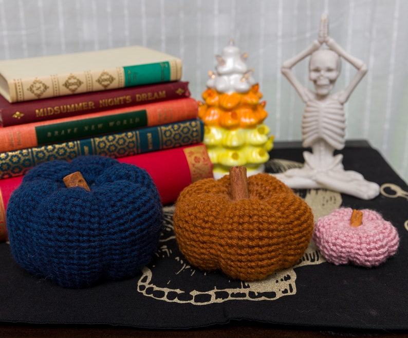 May include: Three crocheted pumpkins in different colors: navy blue, brown, and pink. The pumpkins are sitting on a black surface with a white skull design.