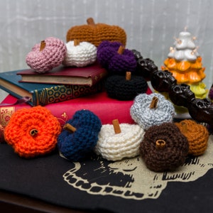 May include: A collection of crocheted pumpkins in various colors, including orange, pink, white, purple, brown, and gray. The pumpkins are arranged on a stack of books and a black surface. Some pumpkins have cinnamon sticks inserted into the top.