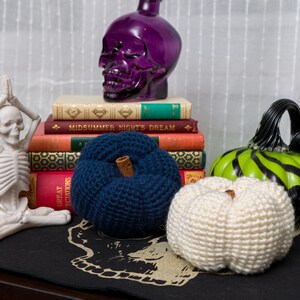 May include: Three crocheted pumpkins, one blue, one white, and one green with black stripes, are arranged on a black surface with a skull design. A purple glass skull sits on top of a stack of books, including one titled "Midsummer Night's Dream."