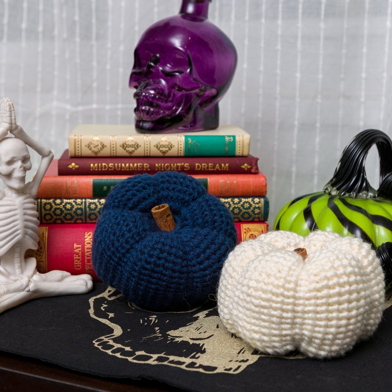 May include: Three crocheted pumpkins, one blue, one white, and one green with black stripes, are arranged on a black surface with a skull design. A purple glass skull sits on top of a stack of books, including one titled "Midsummer Night's Dream."