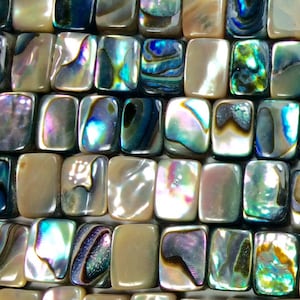 May include: Close-up of a collection of rectangular, iridescent abalone shell beads. The beads display a range of colors, including blues, greens, purples, and creams, with a shimmering, pearlescent effect. The beads are arranged closely together.