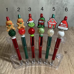 May include: Six red and green glitter pens with different Christmas themed toppers. The toppers include a gingerbread man, a Grinch, a house, and a Santa hat. The pens are displayed in a clear plastic holder.