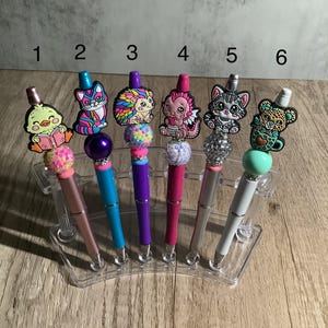 May include: Six colorful pens with different animal toppers. The pens are in a clear plastic holder. The toppers include a duck, a cat, a hedgehog, a dragon, a cat, and a bear.