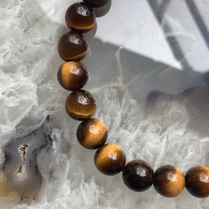 May include: A brown and gold tiger's eye beaded bracelet. The beads are round and smooth, and the bracelet is strung on a clear elastic cord.
