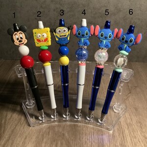 May include: A collection of six decorative pens featuring character toppers. The pens have black, white, and blue barrels. The toppers include Mickey Mouse, SpongeBob SquarePants, a Minion, and three versions of Stitch.