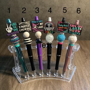 May include: Six decorative pens with different colorful and humorous designs on the tops. The pens are displayed in a clear plastic pen holder.