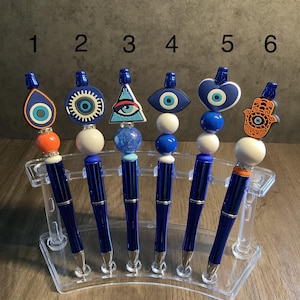 May include: Six blue and white pens with different evil eye charms on the top. The charms include a teardrop, a circle, a triangle, a circle, a heart, and a hand. The pens are displayed on a clear plastic stand.