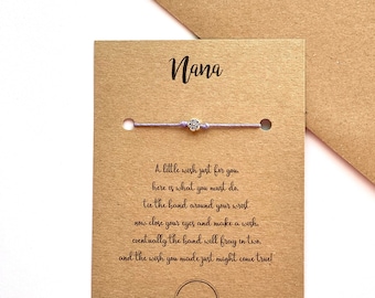 May include: A light purple friendship bracelet with a silver charm in the shape of a flower. The bracelet is displayed on a brown card with the word "Nana" and a message about making a wish.
