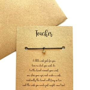 May include: A brown card with the word "Teacher" printed on it. A black string bracelet with a gold star charm is attached to the card. The card includes instructions for making a wish on the bracelet.