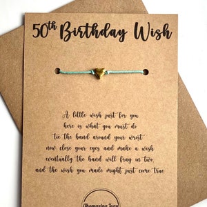 May include: A brown card with the text "50th Birthday Wish" printed in black. A blue string bracelet with a gold heart charm is attached to the card. The card also includes instructions for making a wish on the bracelet.