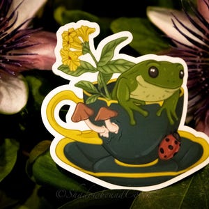 Op de afbeelding: Een groene kikker zit in een blauwe theekop met een gele rand. De theekop staat op een schotel met een gele rand. Er zitten gele bloemen en bruine paddenstoelen in de theekop. Een rode lieveheersbeestje zit op de schotel.