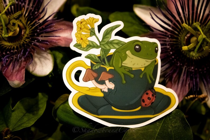 Op de afbeelding: Een groene kikker zit in een blauwe theekop met een gele rand. De theekop staat op een schotel met een gele rand. Er zitten gele bloemen en bruine paddenstoelen in de theekop. Een rode lieveheersbeestje zit op de schotel.