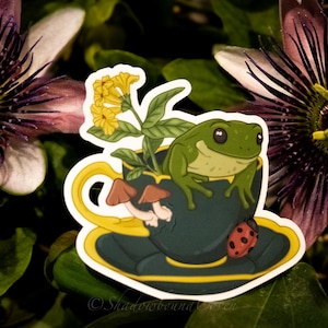 Op de afbeelding: Een groene kikker zit in een blauwe theekop met een gele rand. De theekop staat op een schotel met een gele rand. Er zitten gele bloemen en paddenstoelen in de theekop. Een lieveheersbeestje zit op de schotel. De sticker is op een groene achtergrond.