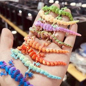 May include: A hand displays a collection of articulated lizard toys in various colours. The toys are in a rainbow of hues, including blue, green, orange, and purple. Each lizard has a segmented body and small legs.
