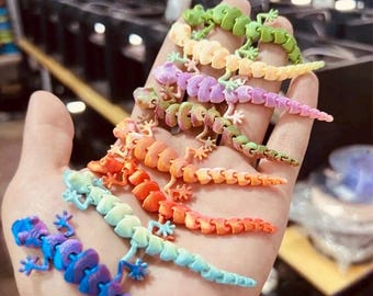 May include: A hand displays a collection of articulated lizard toys in various colours. The toys are in a rainbow of hues, including blue, green, orange, and purple. Each lizard has a segmented body and small legs.