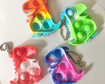 May include: Four dinosaur-shaped silicone sensory fidget keychains. Each toy features a multi-colored tie-dye pattern with circular push-pop bubbles and a silver metal ball chain attached to the top for hanging or securing to personal items.
