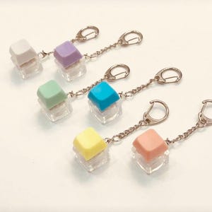 May include: Several keychains with colourful square keycaps on clear, cube-shaped bases. Each keychain has a silver chain and a clasp. The keycaps are in pastel shades: white, purple, green, blue, yellow, and peach.