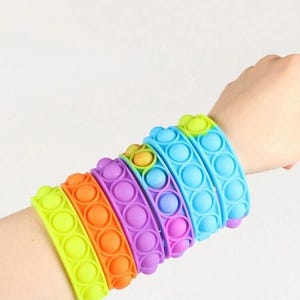 May include: A selection of colourful silicone bubble bracelets in shades of yellow, orange, purple, and blue. Each bracelet has a series of circular bubbles. The bracelets are worn on a wrist, showing the vibrant colours and tactile design.