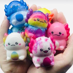 May include: A selection of colourful axolotl figurines in shades of blue, pink, white, and rainbow. Each figure has a smiling face and a unique pattern, held in a person's hands.