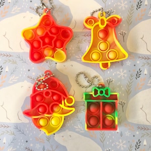 May include: Four colourful Christmas-themed pop it fidget toys with keychains. The toys are shaped like a star, a bell, a Santa hat, and a gift box. The toys are red, yellow, and green.