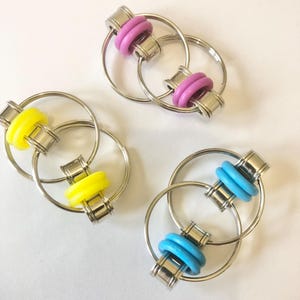 May include: Three metal fidget keychains with colourful rings. The keychains are silver and have two rings each. One keychain has yellow rings, one has pink rings, and one has blue rings.