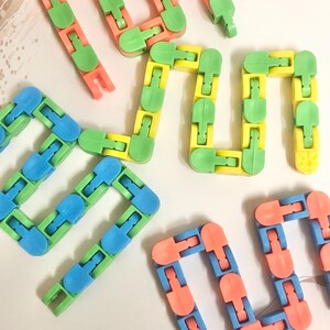 May include: A collection of articulated plastic fidget chains in various colour combinations, including green and orange, green and yellow, and green and blue. Each chain consists of multiple interlocking plastic segments that can be bent.