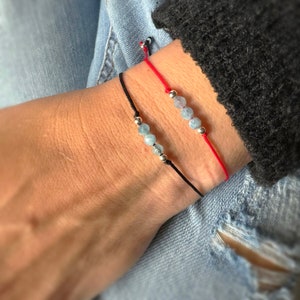 May include: Two bracelets with blue gemstone beads on black and red string. The bracelets are tied around a wrist.