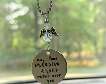 May include: A silver-colored guardian angel car charm. The charm features a small angel with wings and a crystal bead, hanging from a beaded chain. A round metal disc below reads "may Your GUARDIAN ANGEL watch over you."