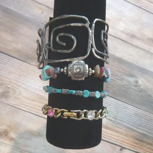 May include: A stack of four bracelets. The top is a wide silver-tone metal cuff with a spiral design. Below it is a beaded bracelet with turquoise stones and a central square silver-tone charm. Next is a turquoise bead bracelet, and the bottom is a gold-tone chain bracelet with inset pink, clear, and green rhinestones.