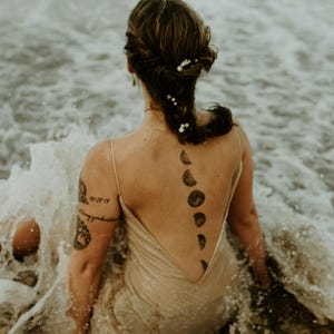 May include: A woman wearing a beige dress sits in the ocean with waves crashing around her. She has a tattoo of a crescent moon on her back, with a series of moons going down her spine.