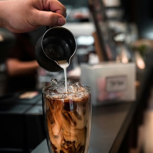 Könnte beinhalten: Ein Glas Eiskaffee, in das Milch gegossen wird. Der Kaffee befindet sich in einem doppelwandigen Glas mit sichtbaren Eiswürfeln. Die Milch wird aus einem kleinen schwarzen Krug gegossen.