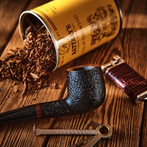 May include: A black pipe with a red band, a yellow tin of "Balkan Sobranie Old & The Cinnamon" tobacco, and a brown leather lighter on a wooden surface.